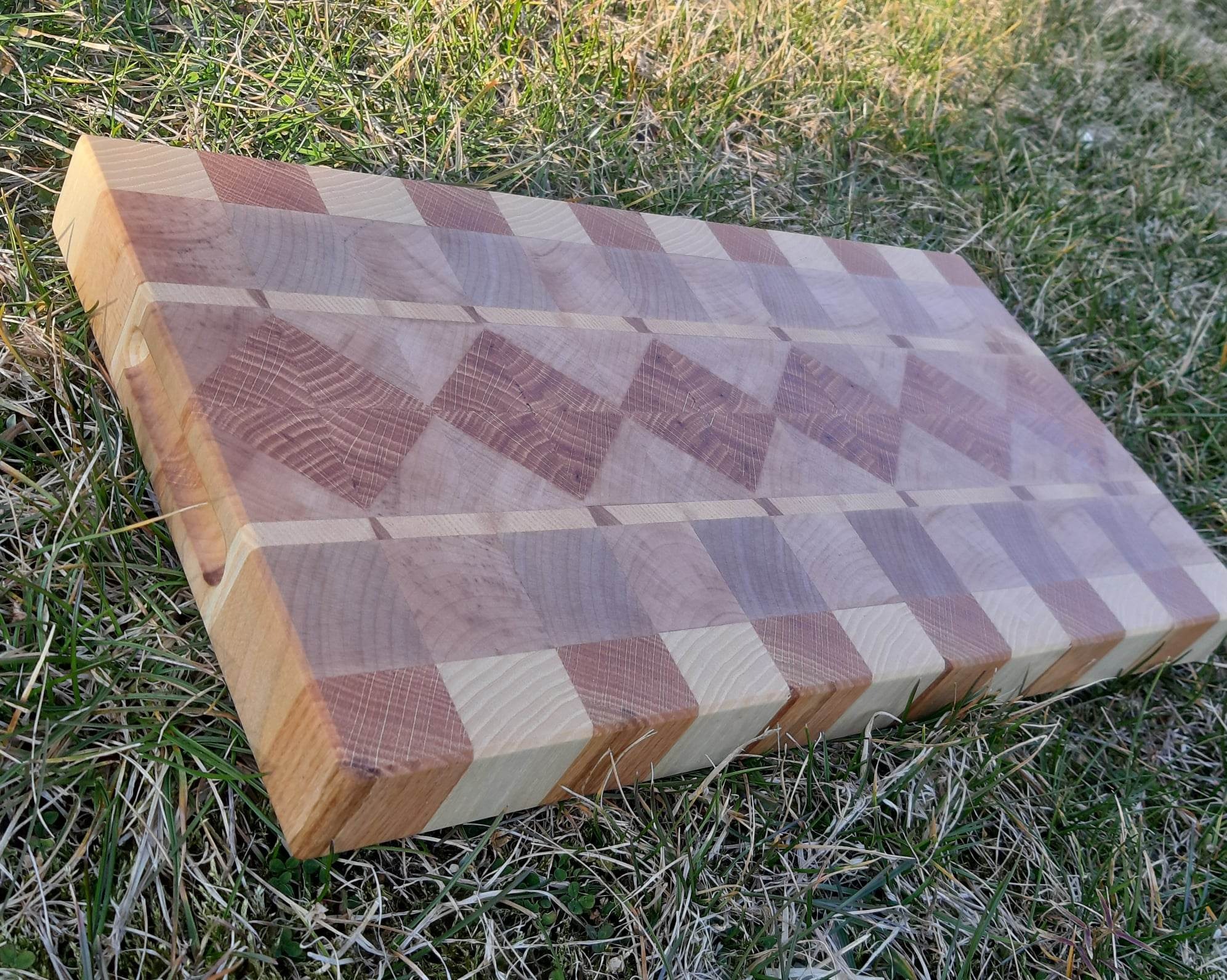 Deska do krojenia z drewna klejonego w geometryczny wzór, leżąca na trawie.