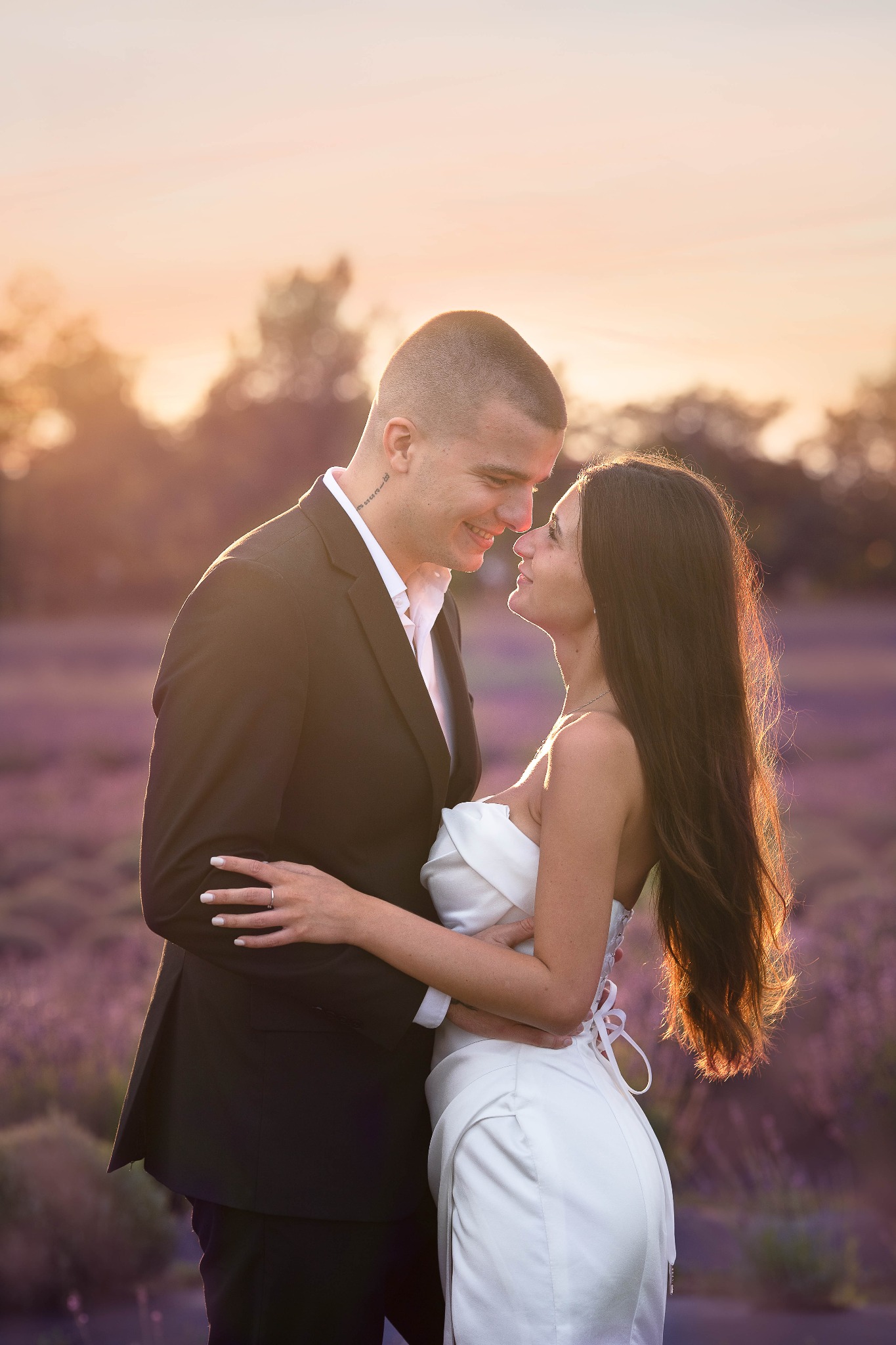 Para młoda, on w czarnym garniturze, ona w białej sukni, obejmująca się na tle fioletowego pola i zachodzącego słońca, z widoczną tatuażą na szyi mężczyzny.