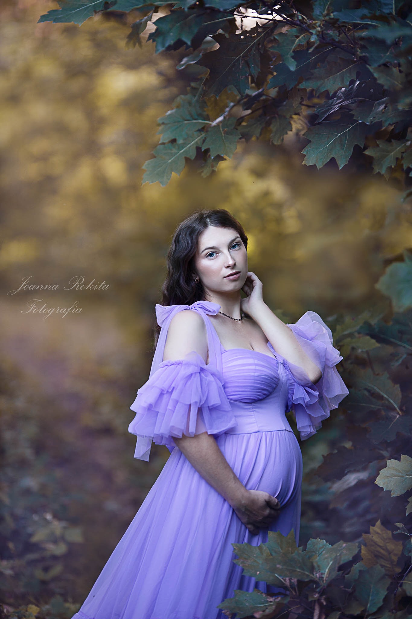 Kobieta w ciąży w fioletowej sukni z falbanami pozuje na tle liści drzew, trzymając dłoń na brzuchu. Delikatne oświetlenie i naturalna sceneria.