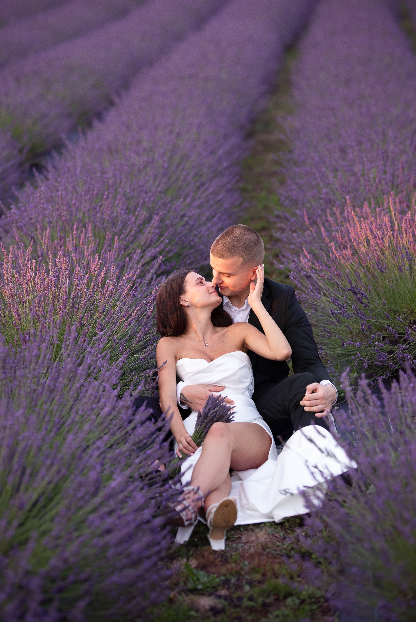 Zakochana para w strojach ślubnych siedzi w polu lawendy, panna młoda trzyma bukiet lawendy, delikatne pocałunek.