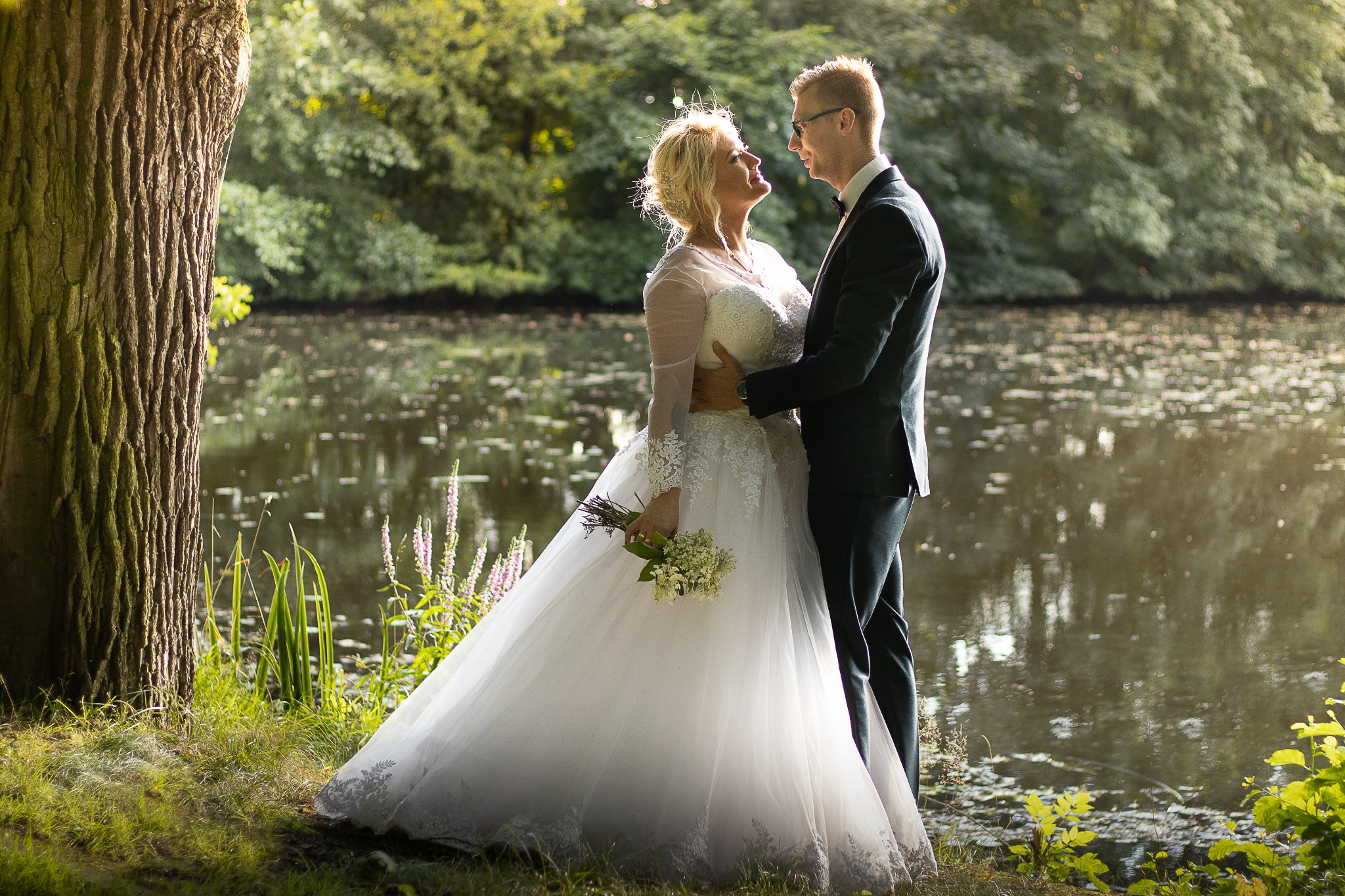 Para młoda w objęciach na tle jeziora i drzew, panna młoda w koronkowej sukni z bukietem, pan młody w czarnym garniturze i okularach.