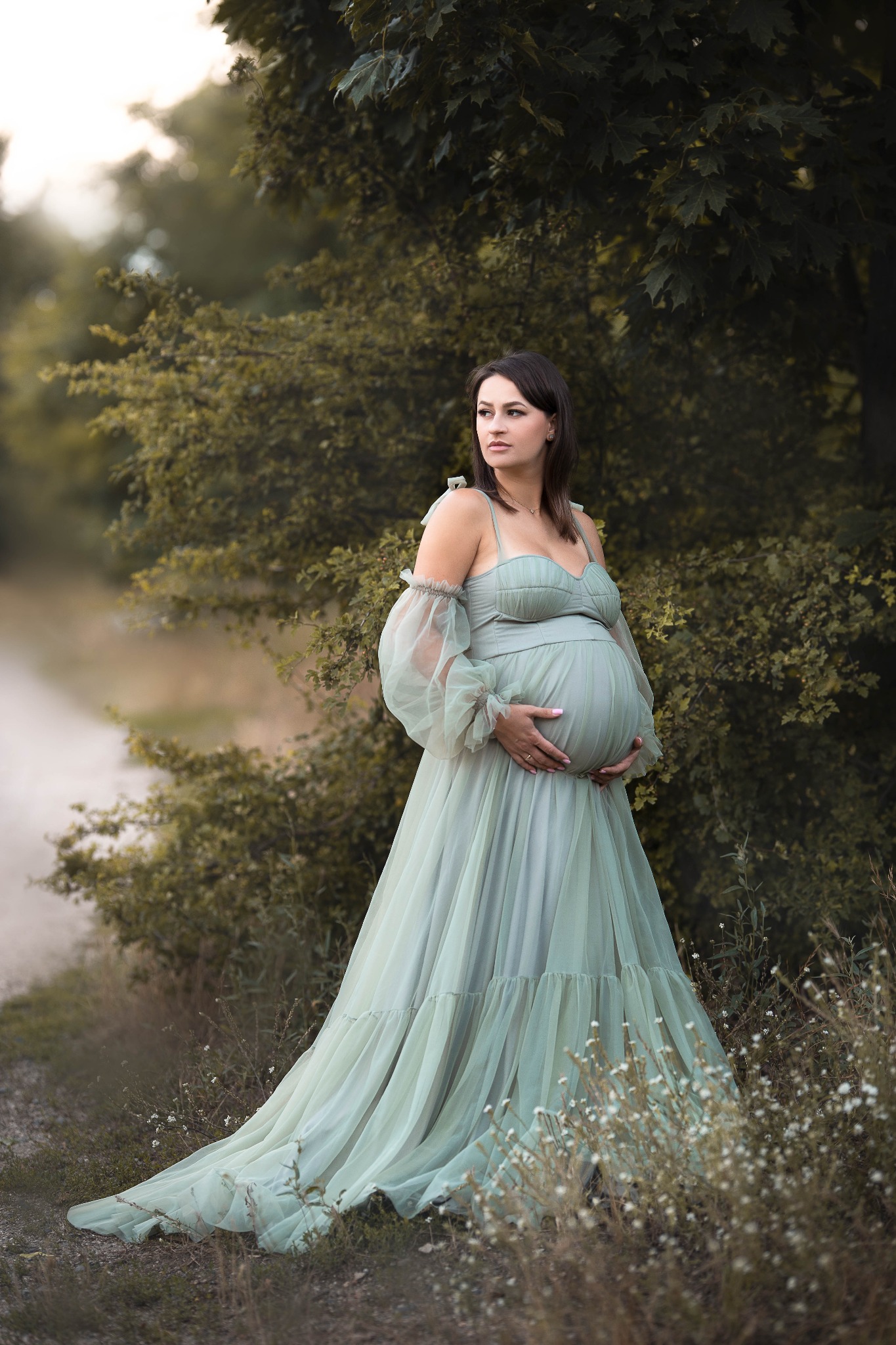 Kobieta w ciąży w długiej, zwiewnej, zielonej sukni z bufiastymi rękawami, pozująca na tle zieleni i polnej drogi, z rękami na brzuchu i wzrokiem skierowanym w lewo.