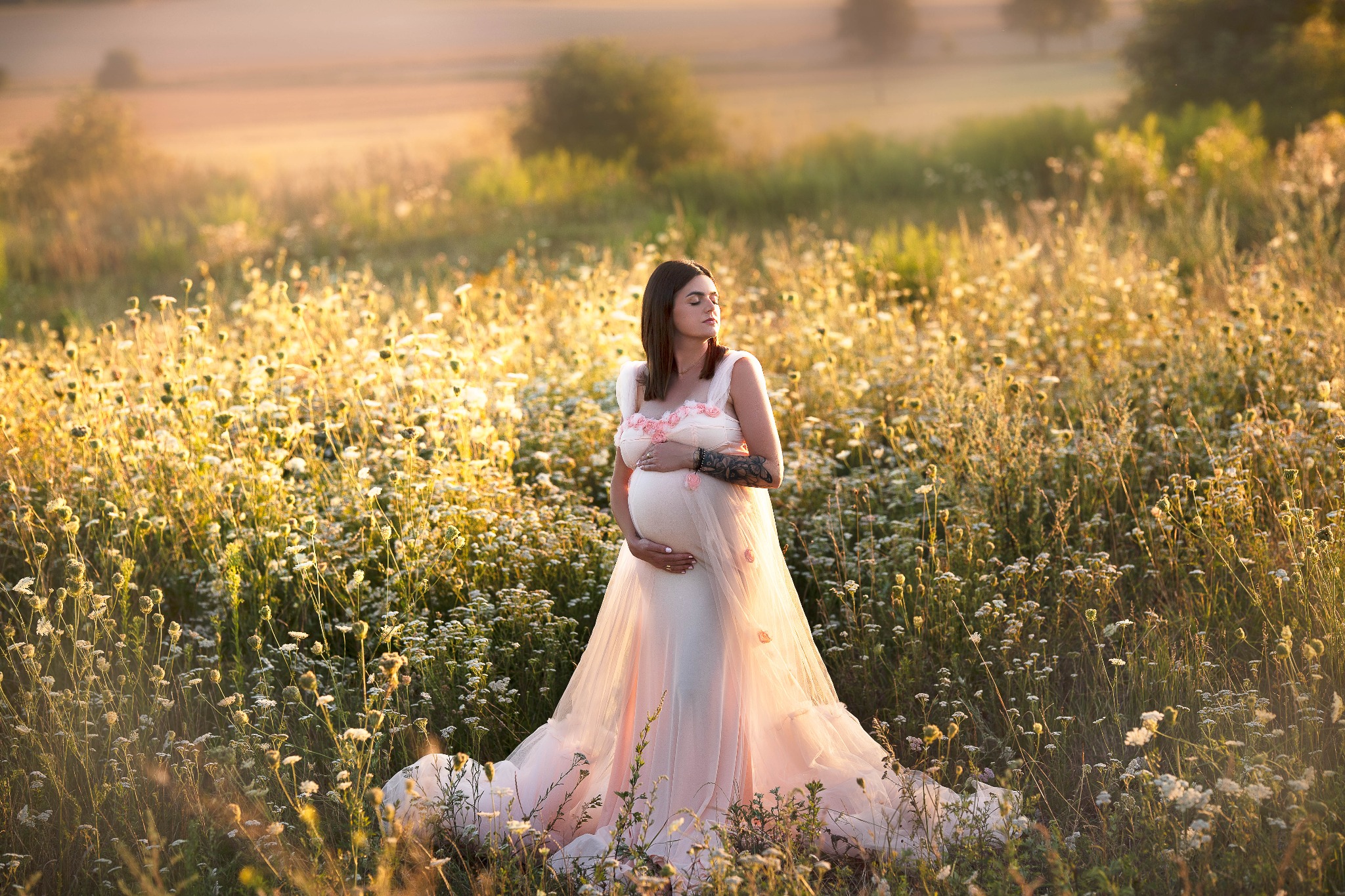 Kobieta w ciąży, w zwiewnej sukni z różowymi aplikacjami, pozuje na łące pełnej kwitnących roślin w promieniach zachodzącego słońca, z zamkniętymi oczami.