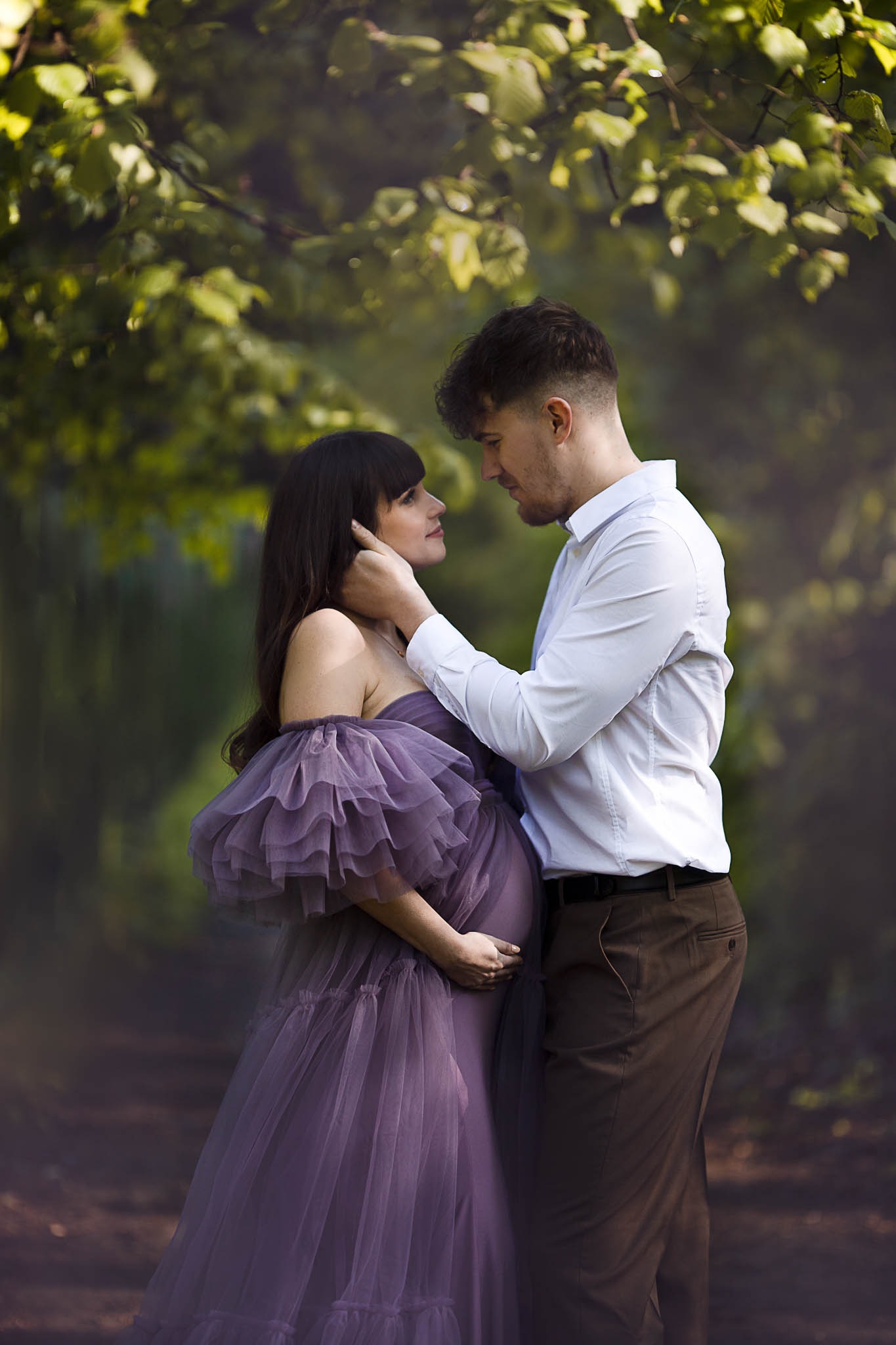 Kobieta w fioletowej, tiulowej sukni ciążowej i mężczyzna w białej koszuli, stojący w zalesionym otoczeniu, patrzący na siebie z czułością.