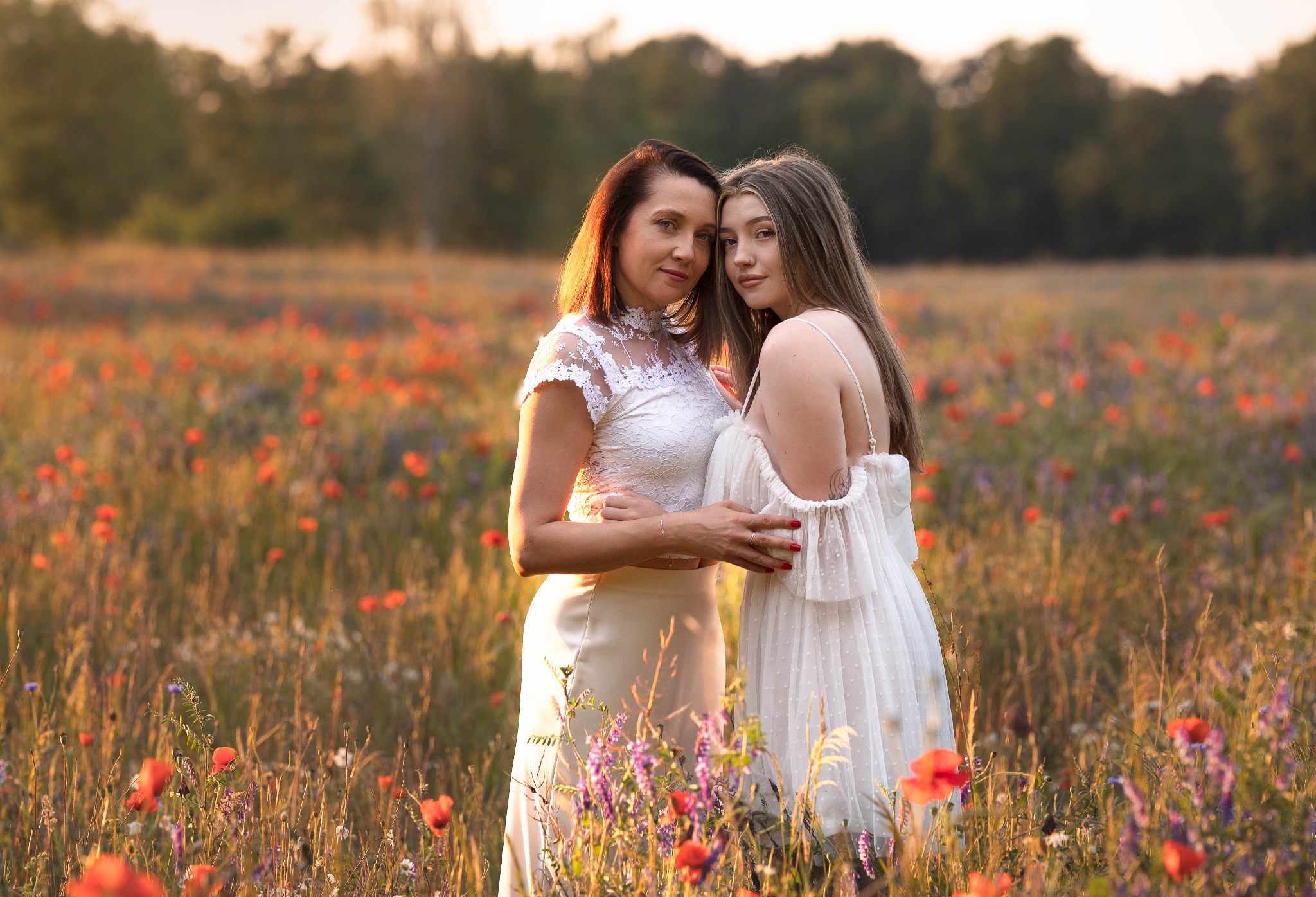 Dwie kobiety, matka i córka, w białych sukienkach, obejmują się na łące pełnej maków i innych polnych kwiatów w ciepłym, zachodzącym słońcu.