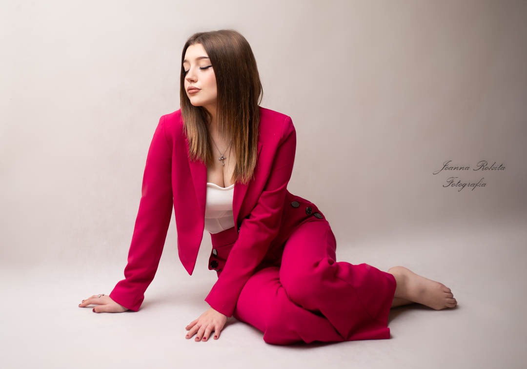 Portret kobiety w malinowym garniturze, pozującej na podłodze w studio, z delikatnym makijażem i zamkniętymi oczami.