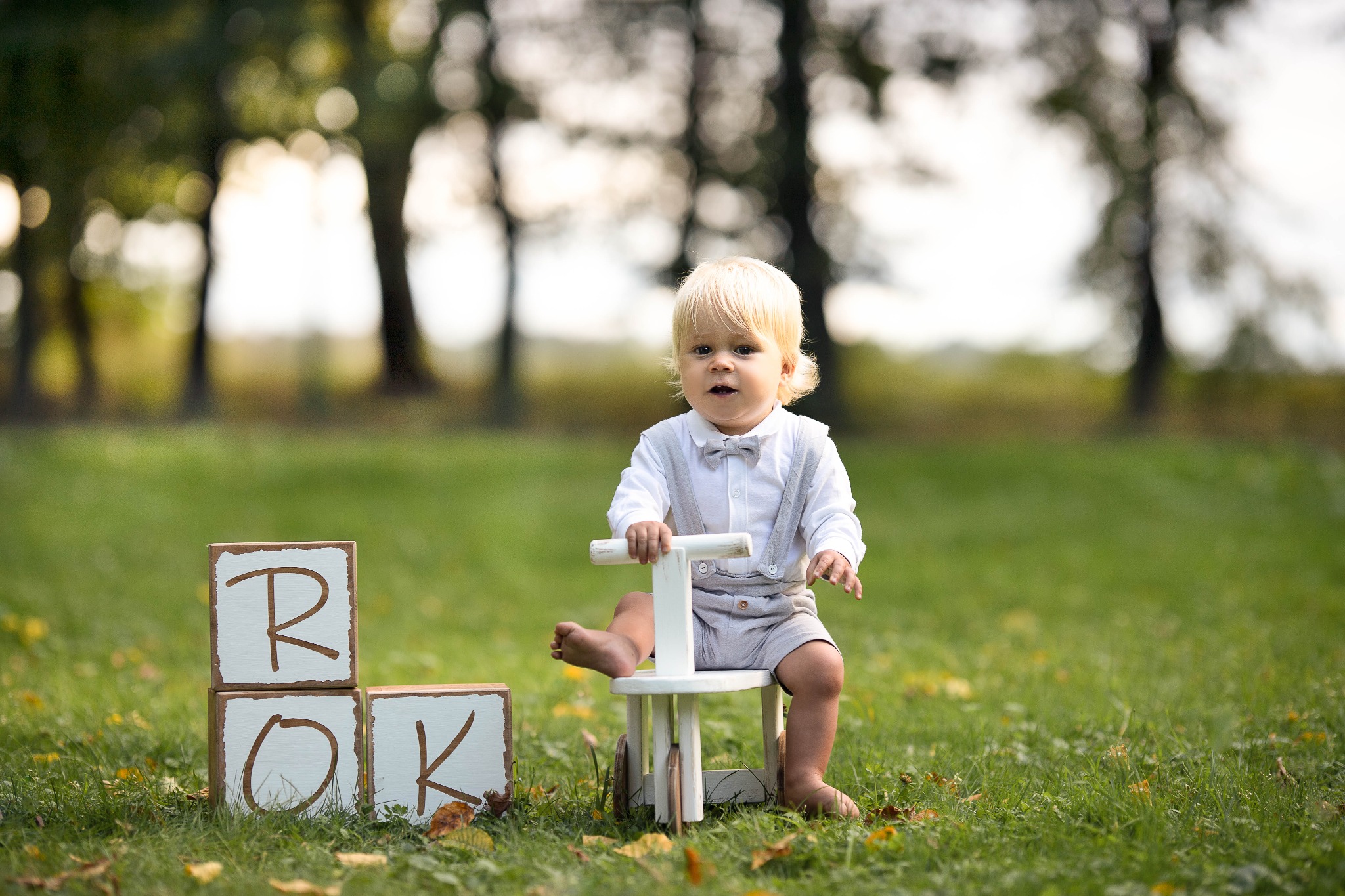 Chłopiec w stroju z muchą siedzi na drewnianym rowerku, obok kostki z literami R, O, K na trawie w parku.
