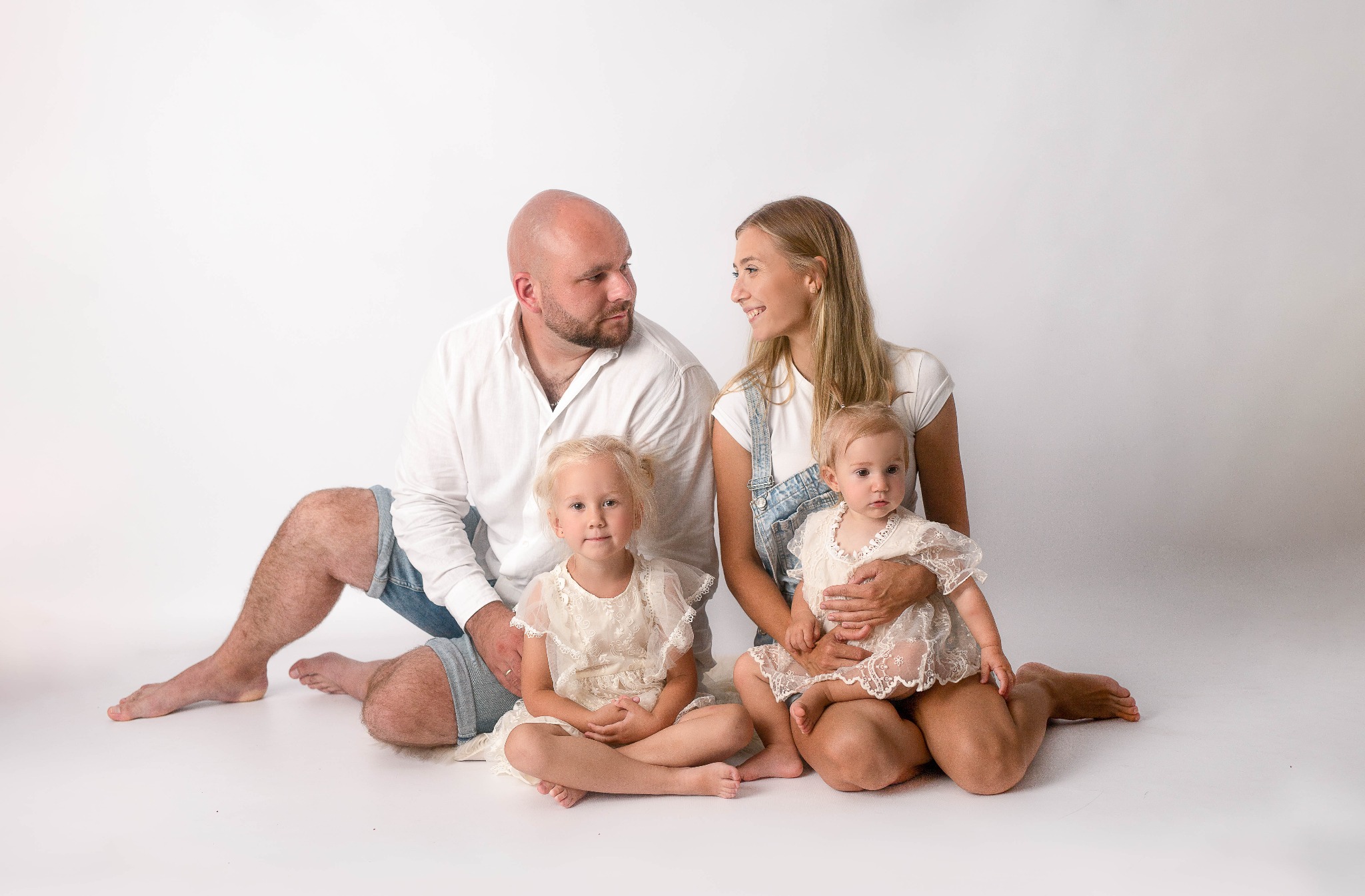 Rodzinne portret studyjny: ojciec w białej koszuli i dżinsowych spodenkach, matka w dżinsowych ogrodniczkach, dwie córki w koronkowych sukienkach siedzą na białym tle.