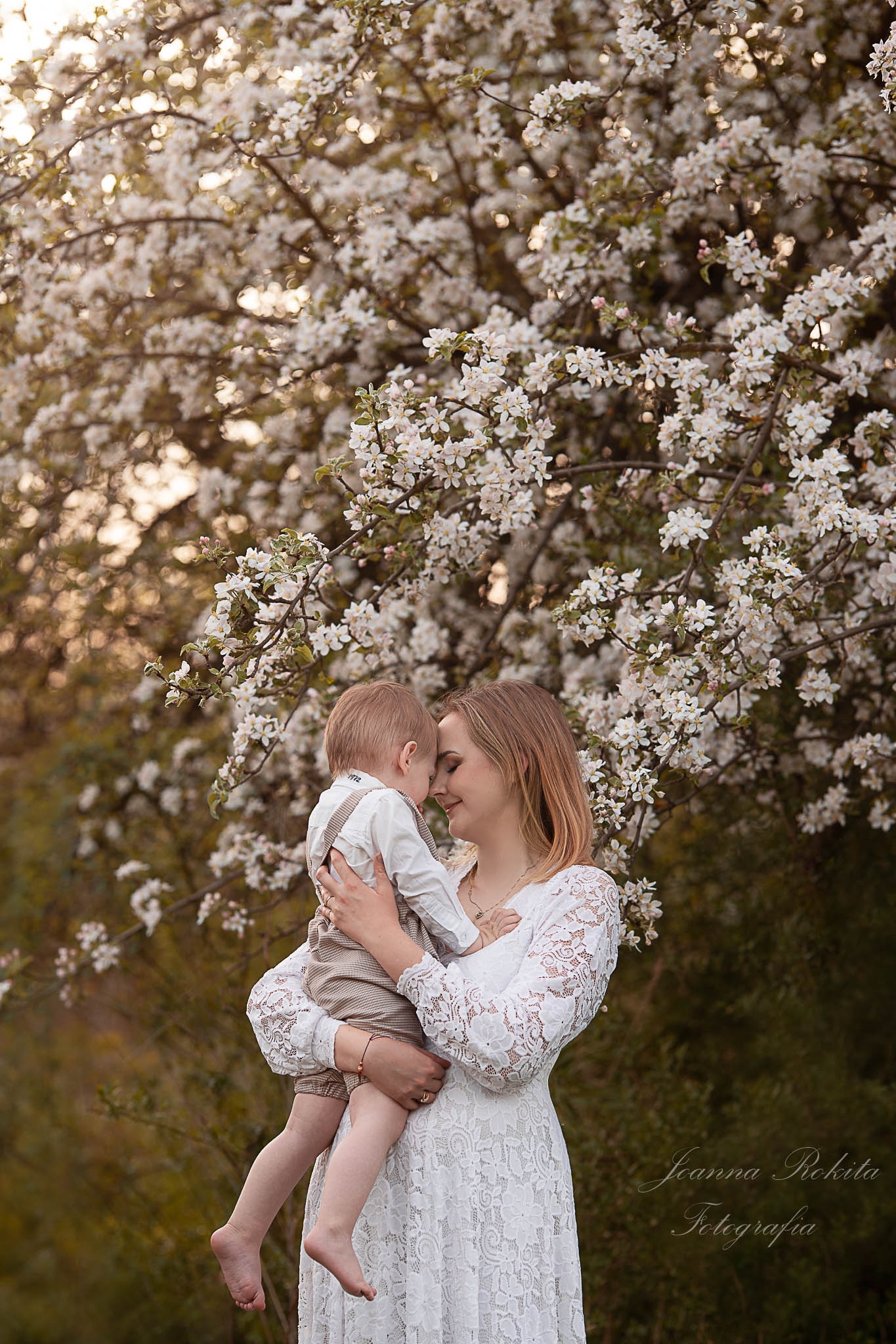 Matka w koronkowej sukience trzyma na rękach małego synka na tle kwitnącego drzewa jabłoni w ciepłym, naturalnym świetle.