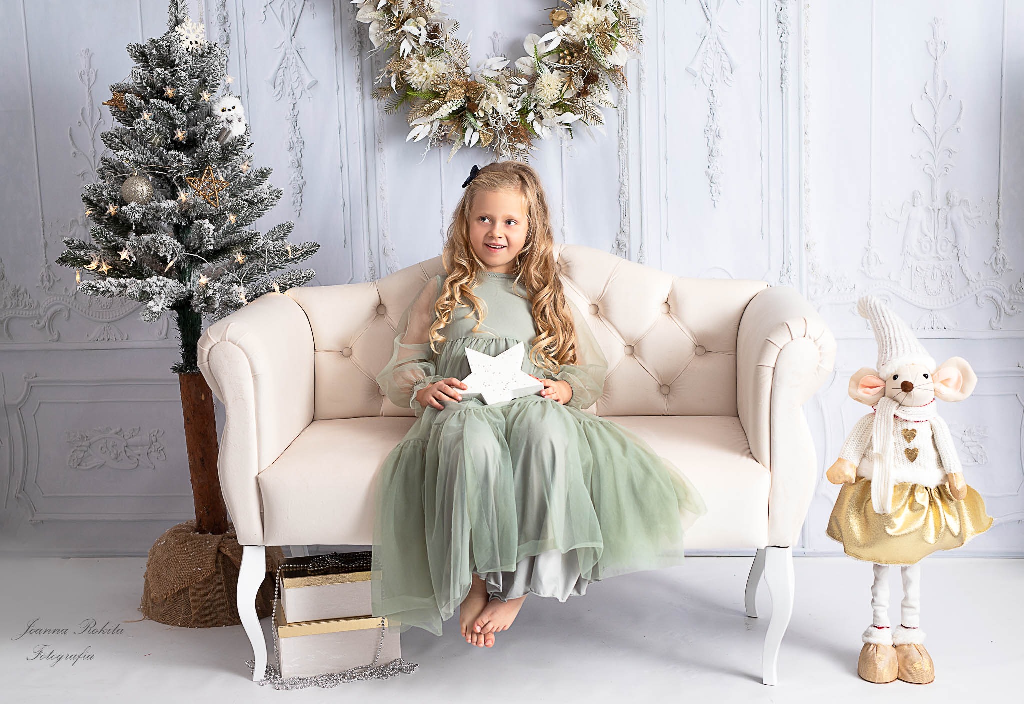 Dziewczynka w tiulowej sukience siedzi na kremowej sofie, trzymając świecącą gwiazdę; obok choinka i pluszowa myszka w zimowej stylizacji, w tle biała ściana z dekoracyjnymi sztukateriami i wieniec.