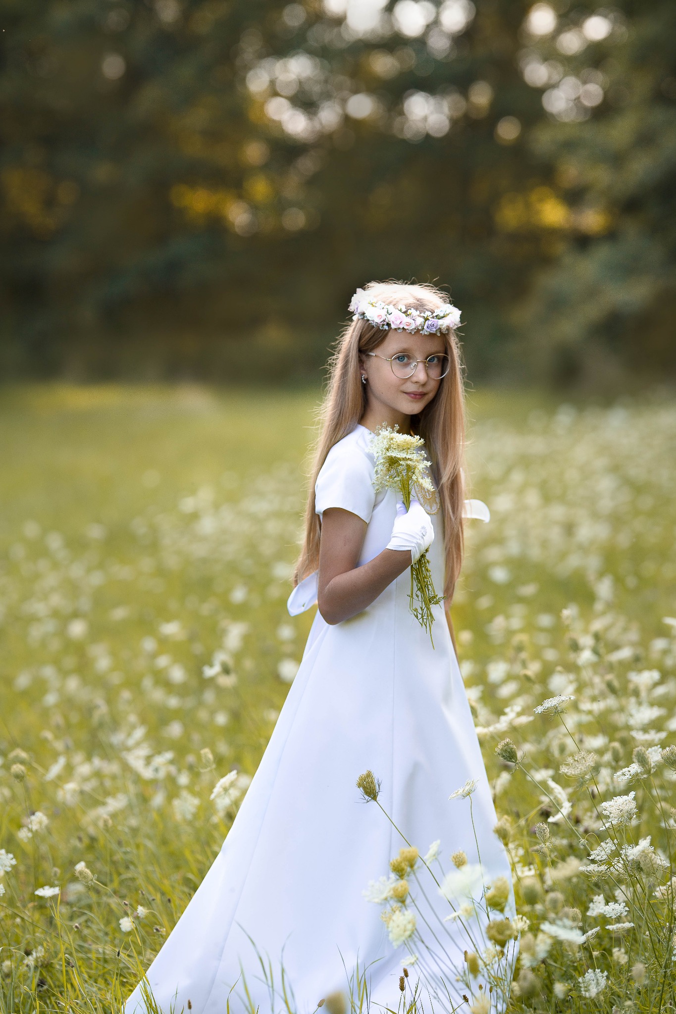 Dziewczynka w białej sukience i wianku na głowie, w okularach, stoi na łące pełnej białych kwiatów, trzymając bukiecik.