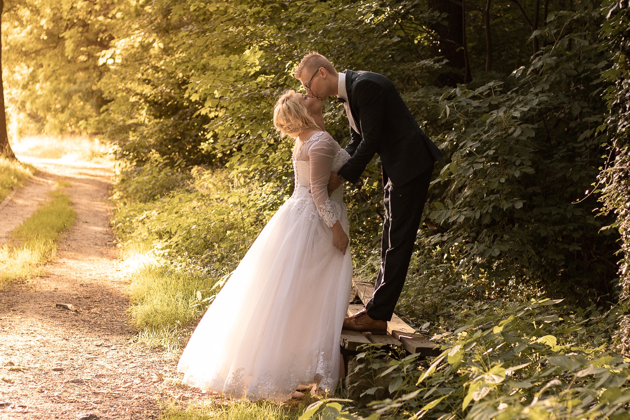 Para młoda całująca się na drewnianym mostku w lesie, panna młoda w koronkowej sukni z długim rękawem, pan młody w ciemnym garniturze i okularach, leśna ścieżka w tle.