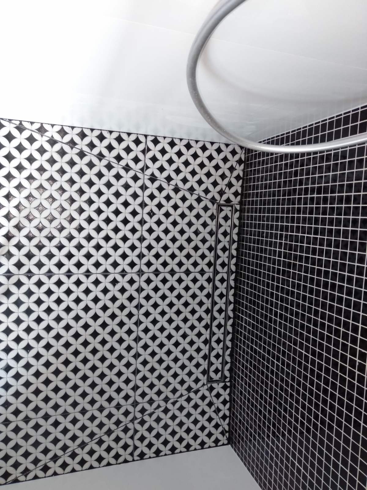 Wykończona ściana łazienki z czarno-białymi płytkami o geometrycznym wzorze i czarnymi, kwadratowymi płytkami z białymi fugami; widoczny odpływ liniowy i fragment chromowanej rury.