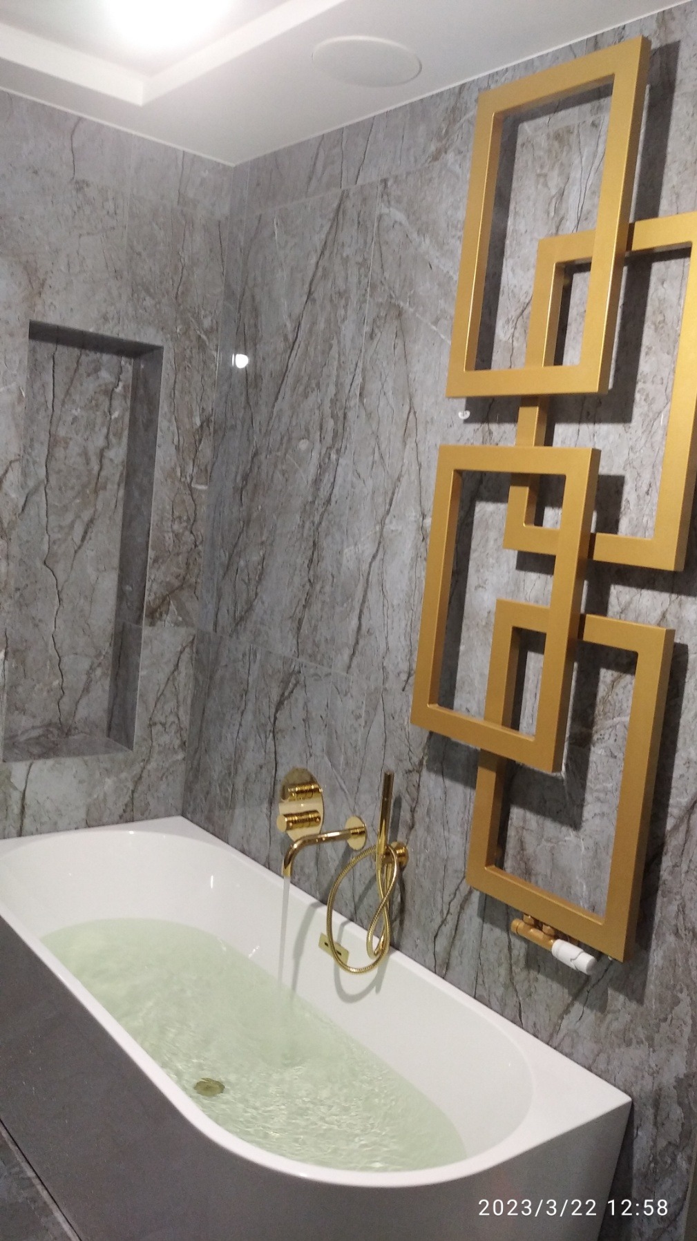 Nowoczesna łazienka z białą wanną wypełnioną wodą, złotą armaturą i dekoracyjnym, złotym grzejnikiem w kształcie połączonych kwadratów na ścianie wyłożonej szarymi płytkami imitującymi marmur.