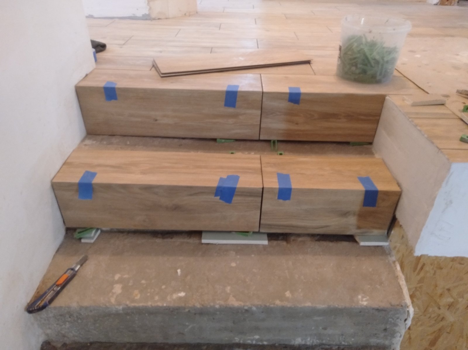 Montaż płytek imitujących drewno na schodach, zabezpieczone niebieską taśmą, widoczne poziomice, nożyk do tapet na dolnym stopniu, etap prac wykończeniowych.