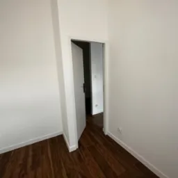 Pusty pokój z białymi ścianami, drewnianą podłogą i otwartymi białymi drzwiami do kolejnego pomieszczenia.