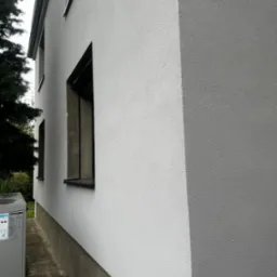 Jasnoszara elewacja budynku z widoczną pompą ciepła i rurami w izolacji, okna z ciemnymi ramami.