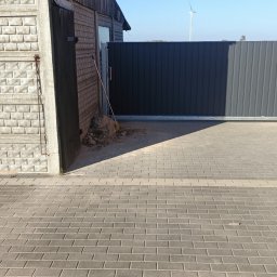 Dammix - Przesuwna brama wjazdowa z ciemnoszarych paneli, widoczny fragment budynku z cegły i drewna oraz sterta ziemi z miotłą, w tle turbina wiatrowa.