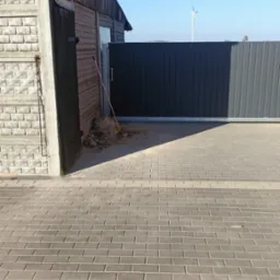 Przesuwna brama wjazdowa z ciemnoszarych paneli, widoczny fragment budynku z cegły i drewna oraz sterta ziemi z miotłą, w tle turbina wiatrowa.