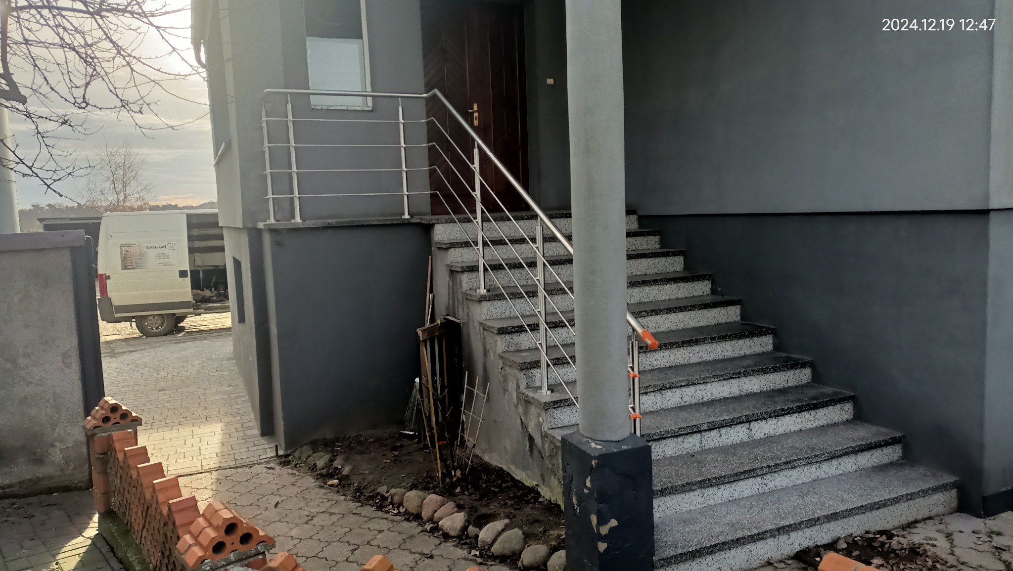 Zewnętrzne schody betonowe z nierdzewną balustradą, prowadzące do wejścia domu o szarej elewacji. Widoczny fragment ceglanego murku, trawnik i samochód dostawczy w tle.