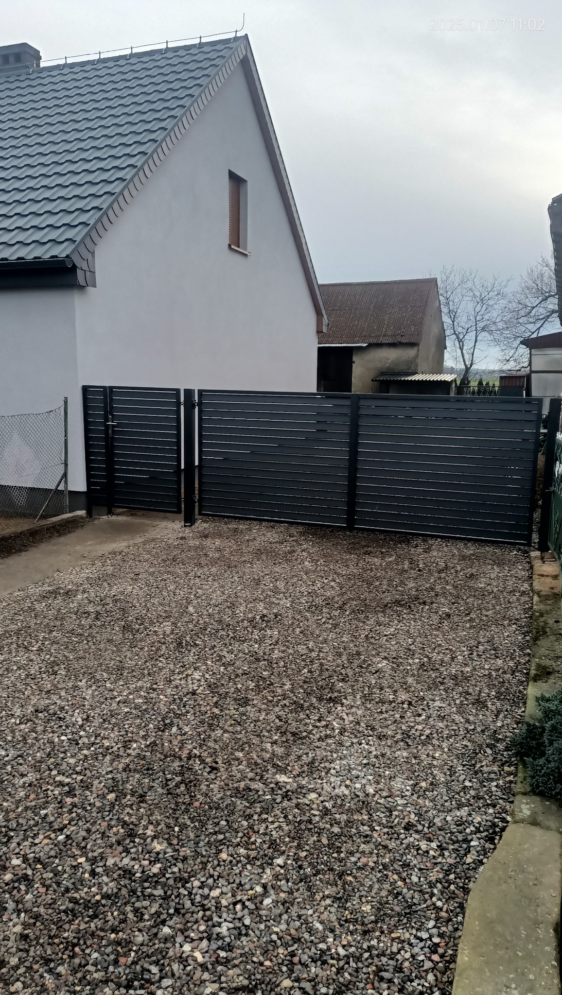 Nowoczesna, pozioma brama wjazdowa z czarnych, wąskich desek, prowadząca na żwirowy podjazd przed domem z szarą dachówką.