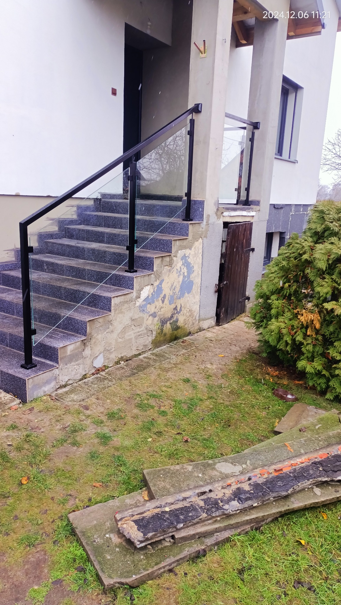 Zewnętrzne schody z szarych płytek z balustradą ze szkła i czarnych metalowych słupków, widoczne niedoskonałości elewacji i fragmenty gruzu na trawniku.