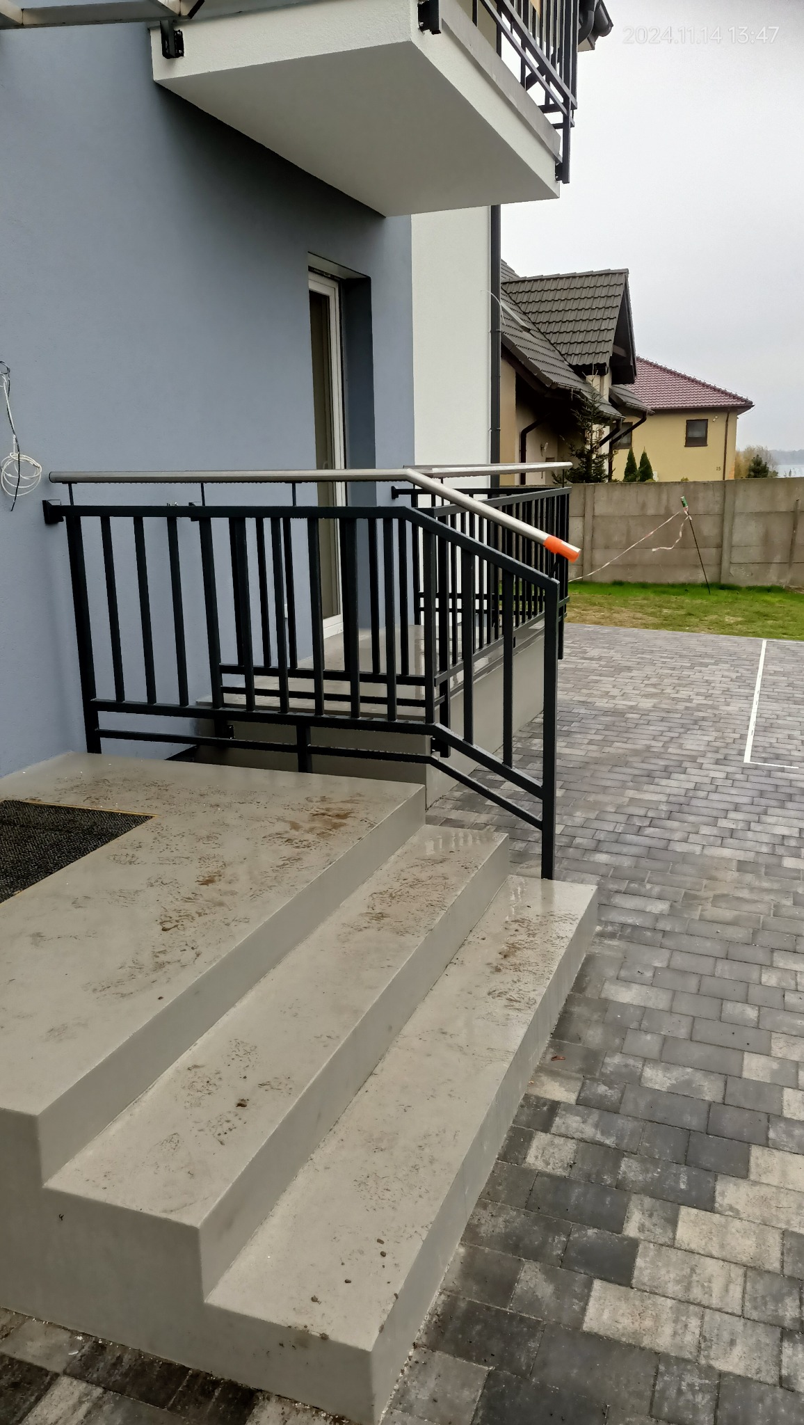 Nowoczesna, czarna balustrada metalowa z poziomymi i pionowymi elementami, zamontowana przy wejściu do domu z szarymi schodami betonowymi i kostką brukową na podjeździe.