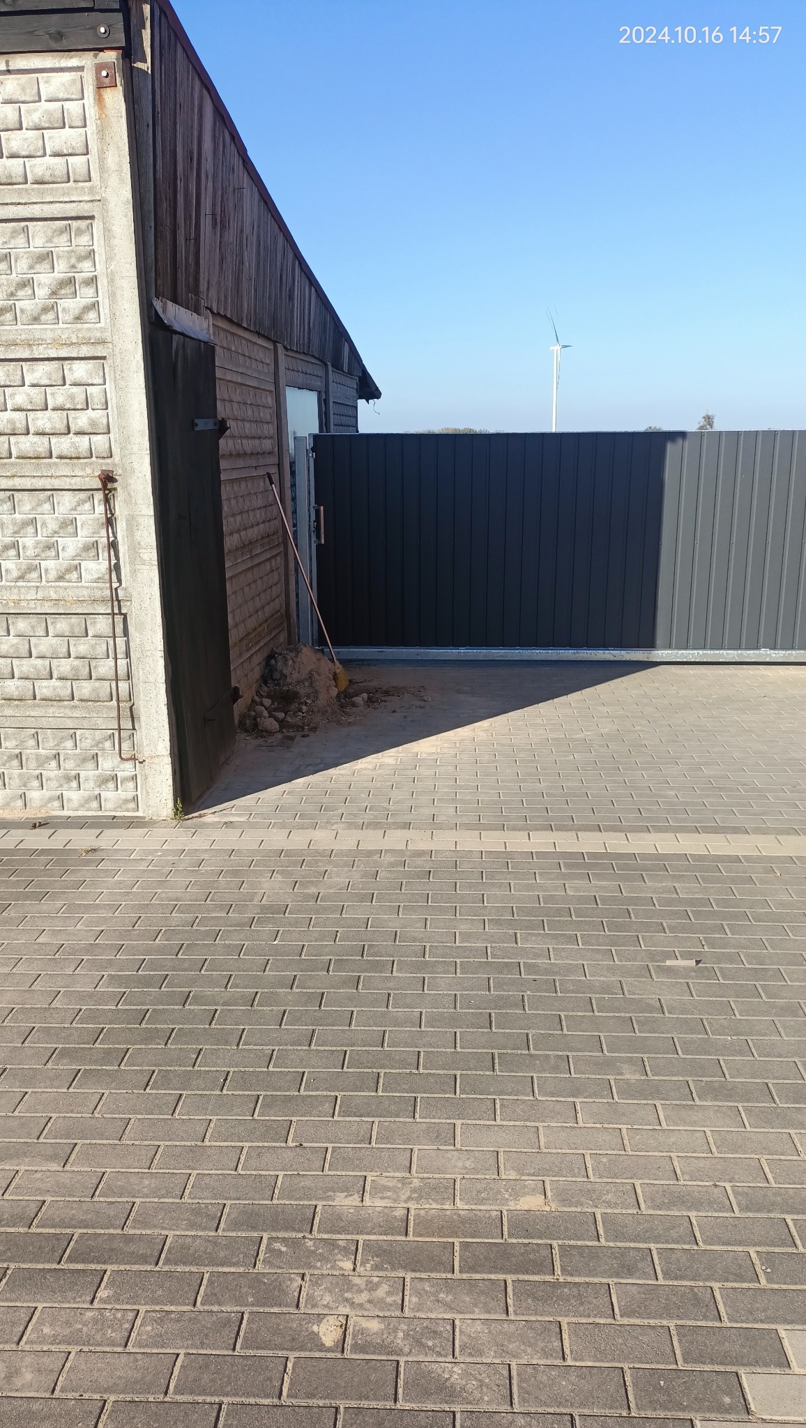Przesuwna brama wjazdowa z ciemnoszarych paneli, widoczny fragment budynku z cegły i drewna oraz sterta ziemi z miotłą, w tle turbina wiatrowa.