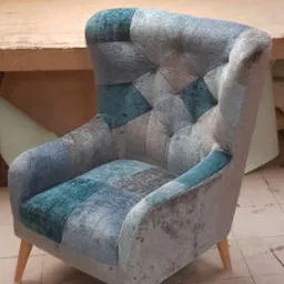 Fotel uszak z patchworkową tapicerką w odcieniach szarości i turkusu, stojący na kafelkach w warsztacie tapicerskim.