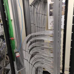 Szczegółowe ujęcie uporządkowanych kabli w szafie serwerowej, z widocznymi wiązkami szarych kabli spiętych czarnymi opaskami, oraz zielonych i niebieskich kabli po lewej stronie, a także elementami...