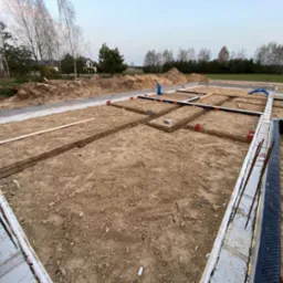 Fundamenty domu w trakcie budowy z widocznymi instalacjami hydraulicznymi, zbrojeniami i izolacją termiczną, w tle zabudowa jednorodzinna i drzewa.