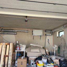 Usługi Hydrauliczne Kamil Borkowski - Montaż instalacji wentylacyjnej z rekuperacją w garażu. Widoczne rury wentylacyjne, centrala rekuperacyjna i bałagan po montażu. Ujęcie z dołu, widoczne elementy konstrukcyjne sufitu.