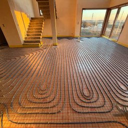 Usługi Hydrauliczne Kamil Borkowski - Ułożone rury ogrzewania podłogowego na czerwonej macie w jasnym pomieszczeniu z oknem i schodami. Widoczne elementy instalacji i przygotowanie do wylewki.