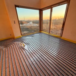 Usługi Hydrauliczne Kamil Borkowski - Ułożone rury ogrzewania podłogowego w jasnym pomieszczeniu z dużym oknem, widok na zewnątrz oświetlony zachodzącym słońcem. Żółta taśma zabezpieczająca wzdłuż ścian.