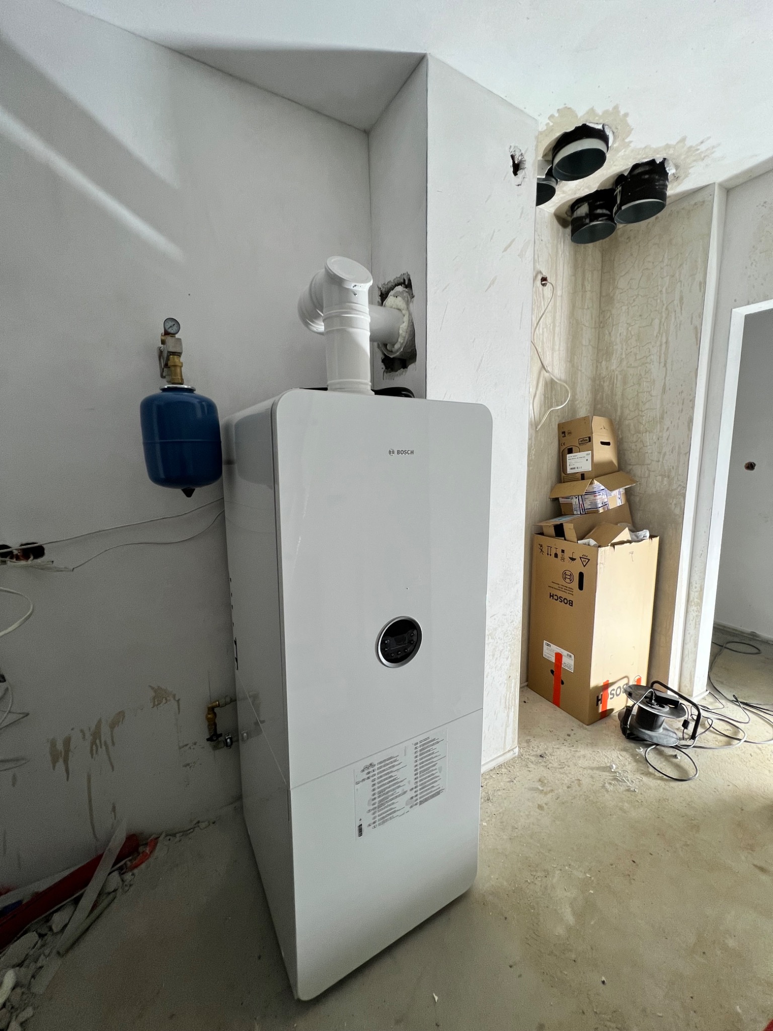 Biały piec gazowy marki Bosch, z podłączonym przewodem kominowym i niebieskim naczyniem wzbiorczym, zamontowany w surowym wnętrzu, widoczne przewody, kartony i otwory wentylacyjne w suficie.
