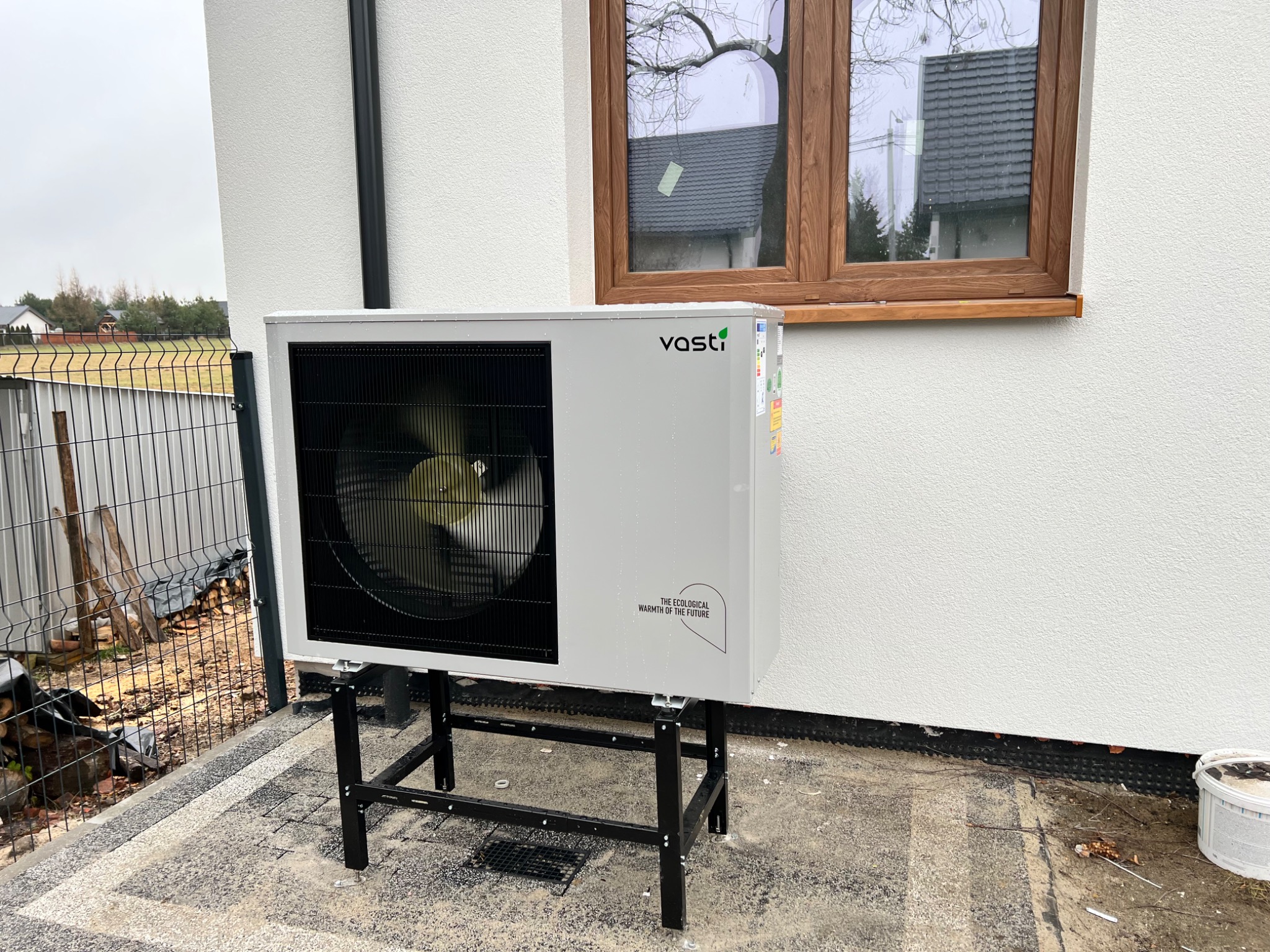 Zewnętrzna jednostka pompy ciepła marki Vasti na czarnym metalowym stelażu, zainstalowana przy białej ścianie domu z drewnianym oknem, widoczny fragment posesji.