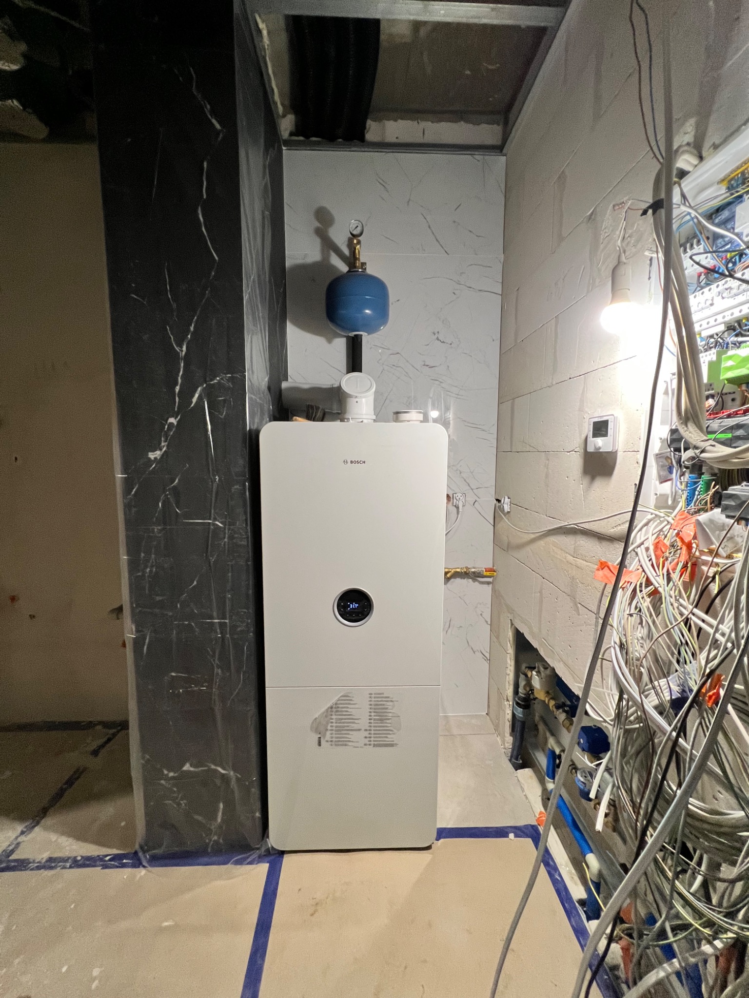 Nowoczesny, biały piec gazowy marki Bosch zamontowany w narożniku pomieszczenia w trakcie prac wykończeniowych, widoczne przewody i nieotynkowane ściany.