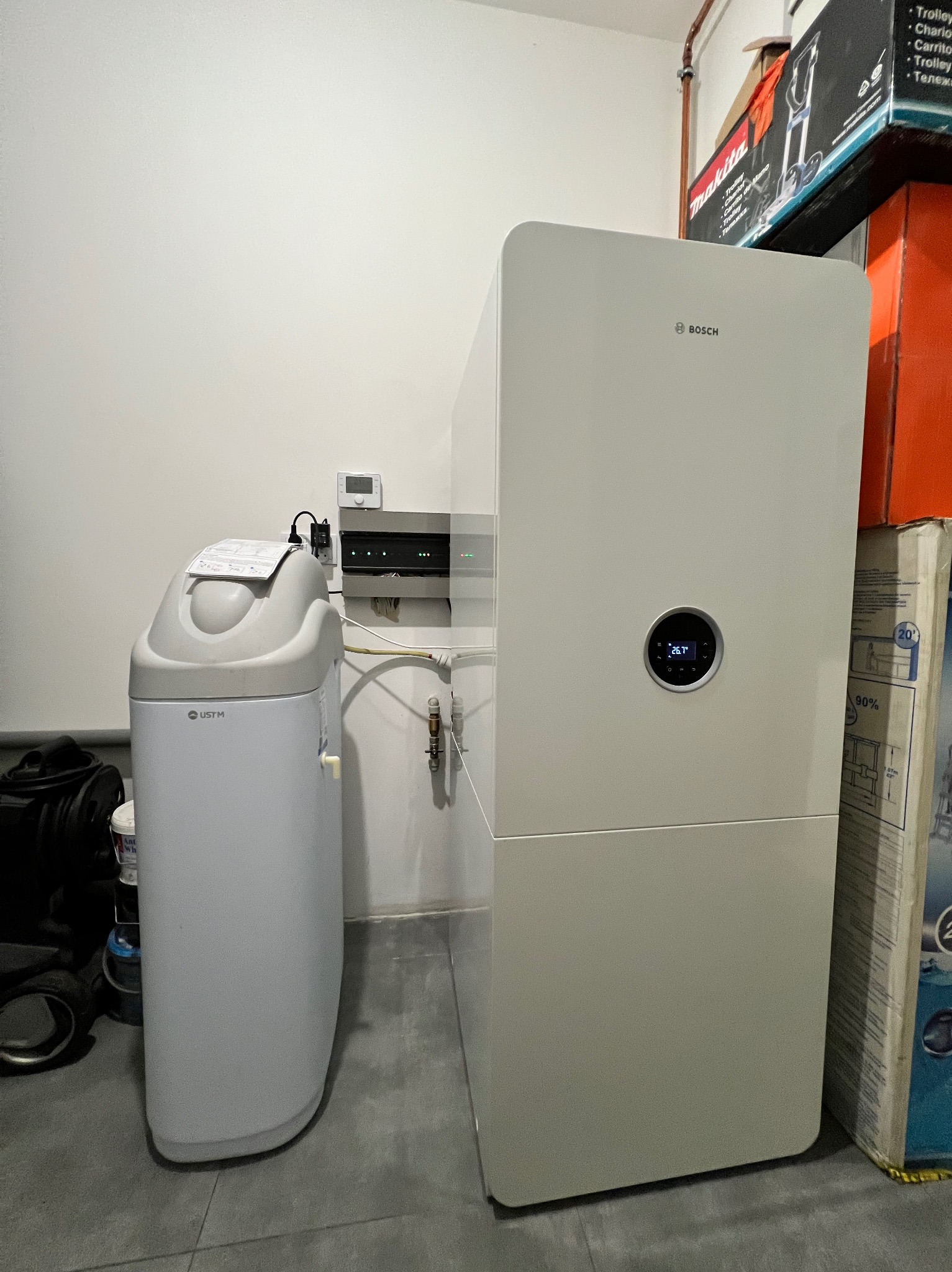 Nowoczesny, biały piec gazowy Bosch z wyświetlaczem temperatury 26.7 stopni Celsjusza, obok zmiękczacz wody UST M, na tle białej ściany i kartonów.