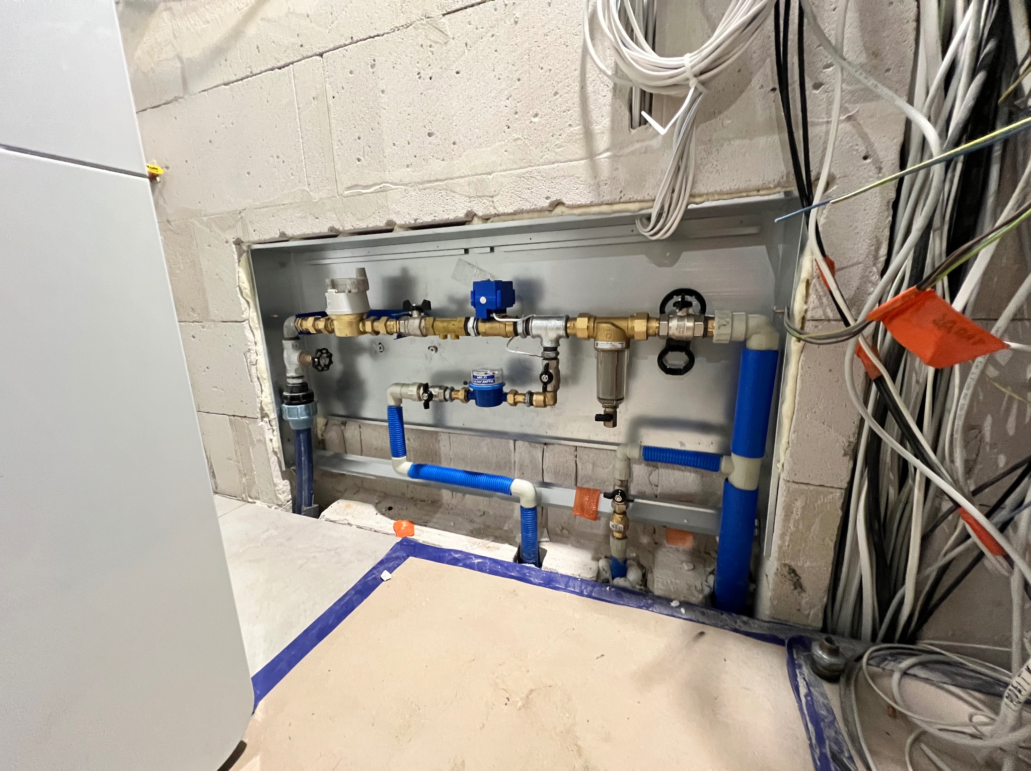 Wnęka z zamontowaną instalacją wodną: rury z niebieskim oplotem, liczniki, zawory, filtry. Widoczne przewody elektryczne i surowa ściana z betonu komórkowego.