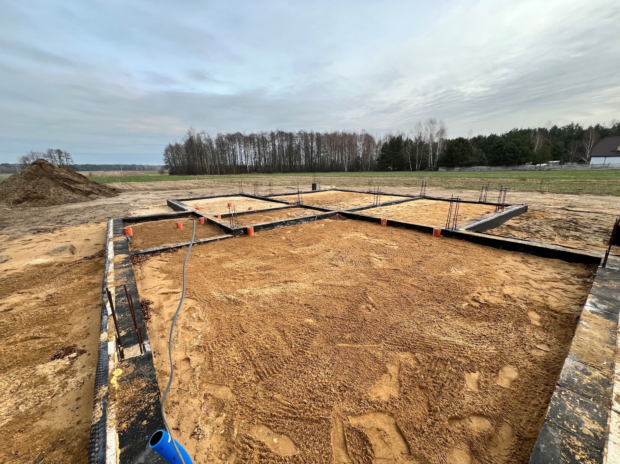 Przygotowany teren pod fundamenty domu jednorodzinnego z widocznymi zbrojeniami i elementami instalacji w gruncie, z kopcem ziemi w tle.