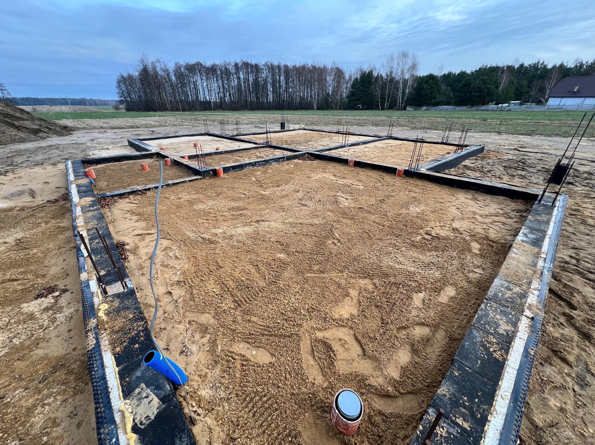 Przygotowany fundament pod budynek jednorodzinny: szalunki zbrojeniowe wypełnione piaskiem, widoczne pionowe pręty zbrojeniowe, niebieska rura i puszka instalacyjna w rogu, tło z drzewami i polem...