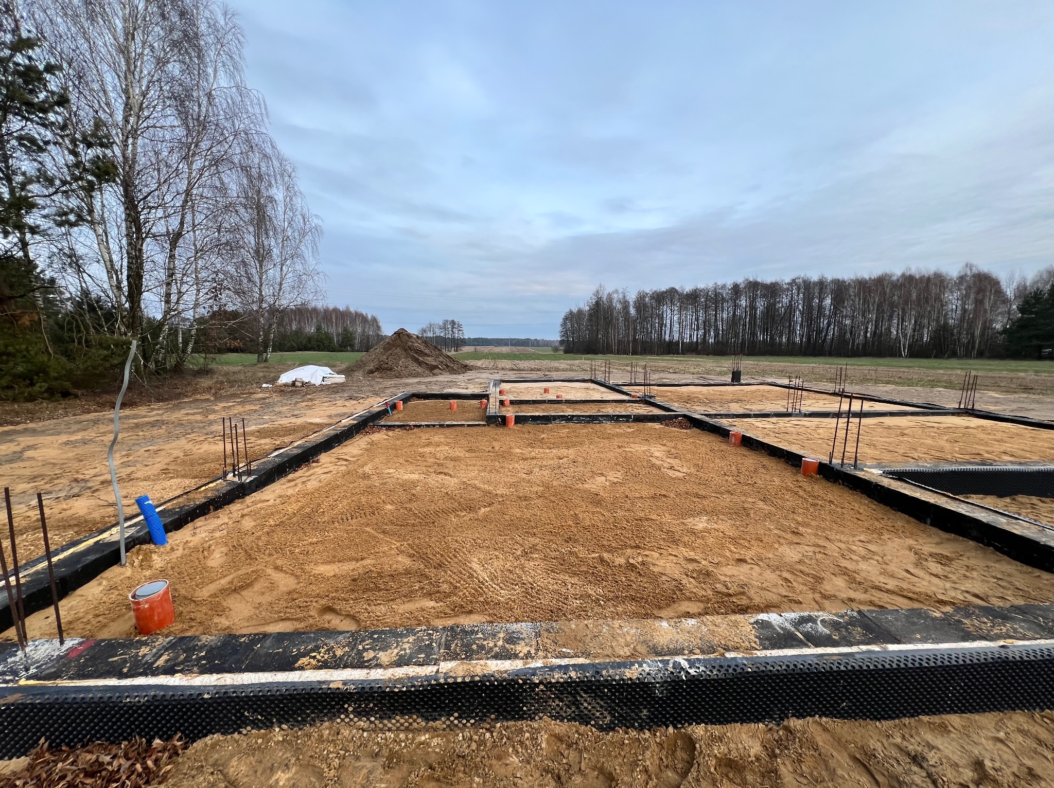 Fundamenty domu jednorodzinnego w Wiśniewie, widok na zbrojenie, izolację przeciwwilgociową i piaskowe wypełnienie, w tle drzewa i pochmurne niebo.