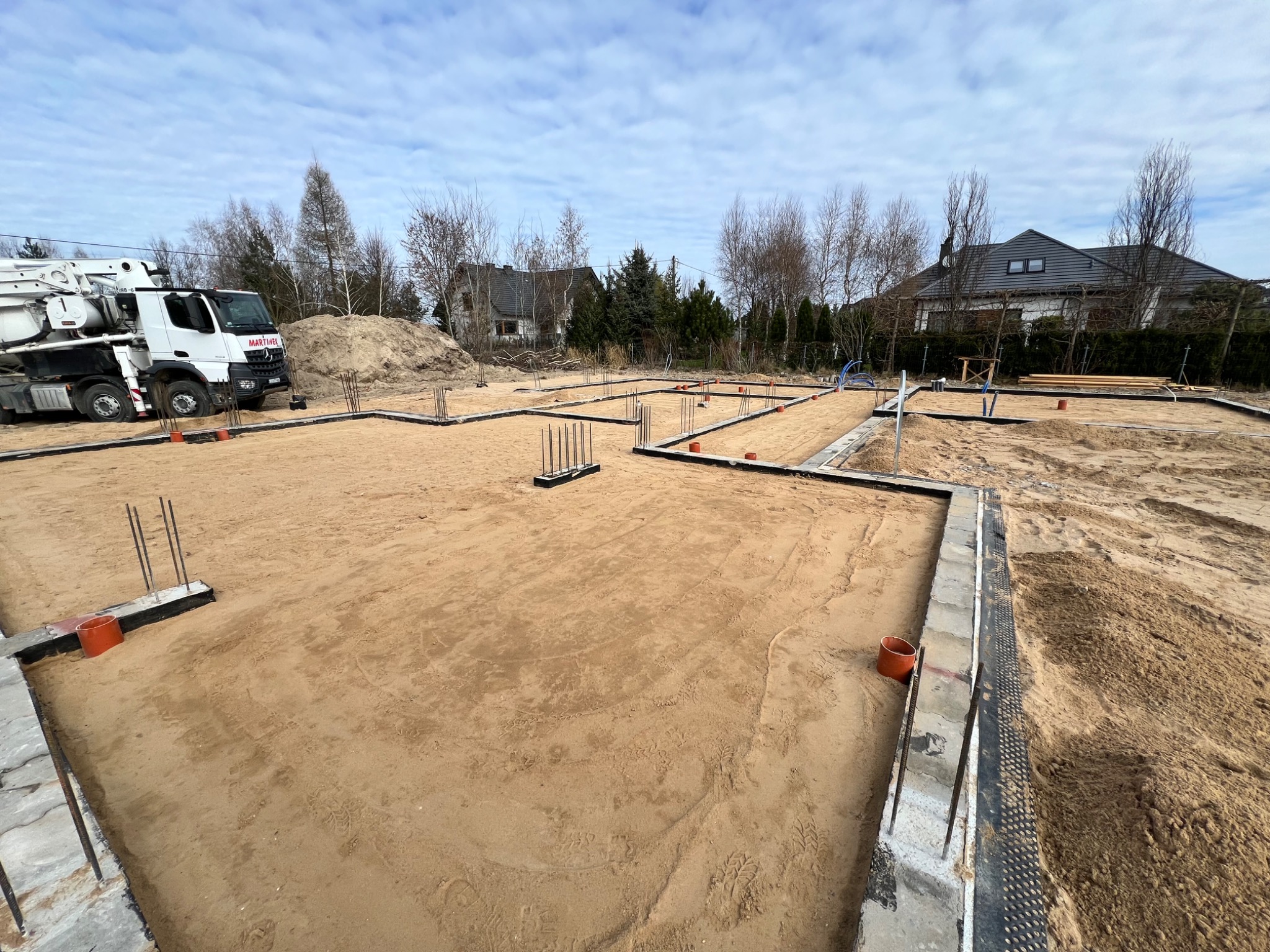 Wykop pod fundamenty domu jednorodzinnego, widoczne zbrojenia, szalunki i pompa do betonu na działce budowlanej w słoneczny dzień.