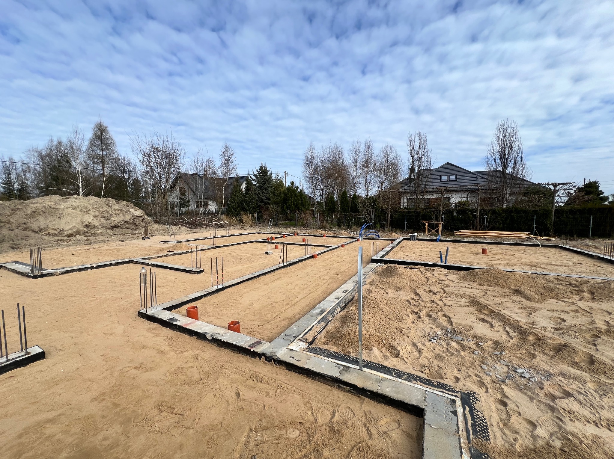 Fundamenty domu jednorodzinnego w trakcie budowy, widoczne zbrojenia i rury instalacyjne, piasek wypełniający przestrzeń między ławami fundamentowymi.