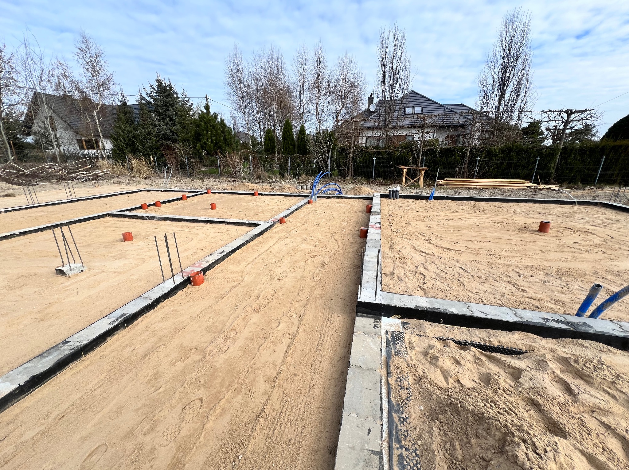 Fundamenty domu w trakcie budowy z widocznymi zbrojeniami, izolacją przeciwwilgociową i rurami instalacji podposadzkowej, na piaszczystym podłożu, z domami w tle pod błękitnym niebem.