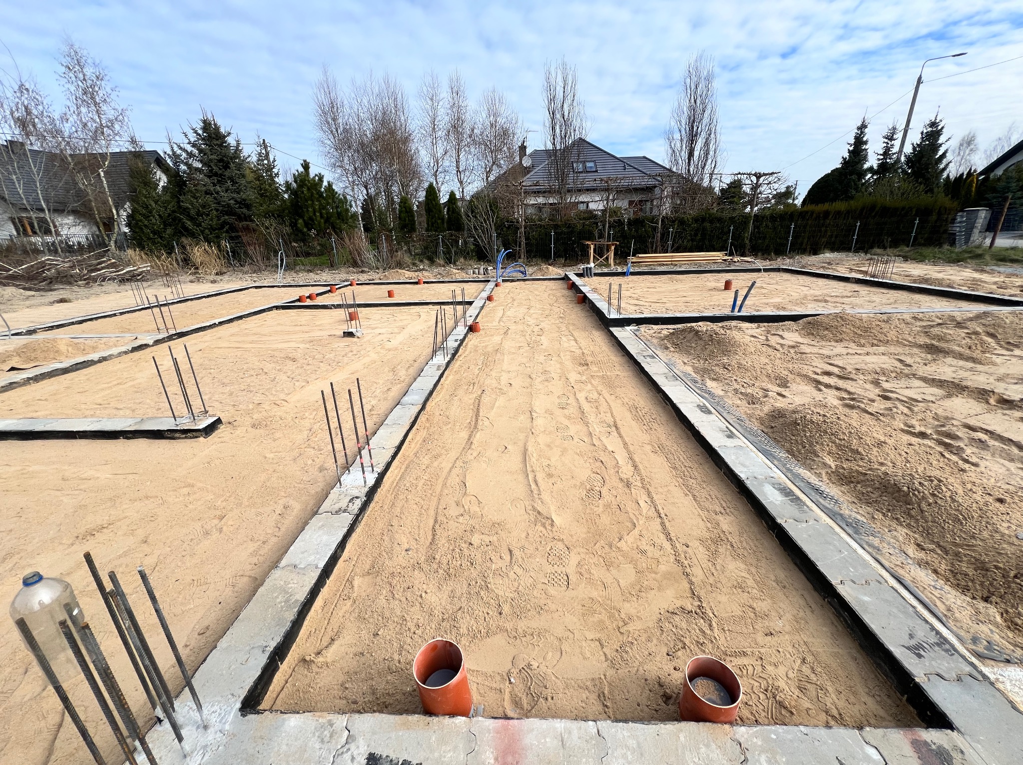 Fundamenty domu jednorodzinnego w trakcie budowy, widoczne zbrojenia, piasek zasypowy, rury instalacyjne wystające z ziemi i kształtki kanalizacyjne w wykopie.