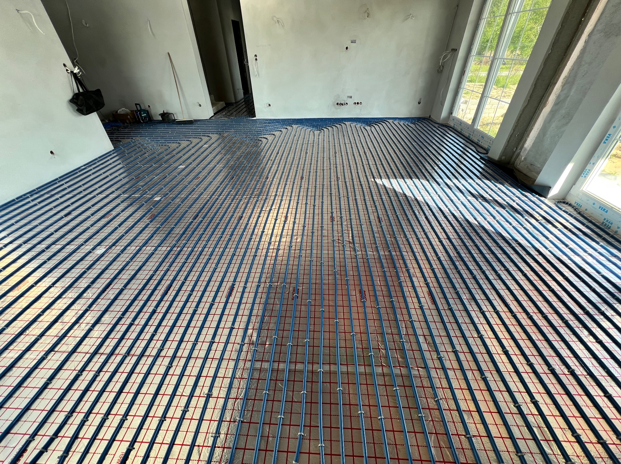 Niebieskie rury ogrzewania podłogowego ułożone na folii z czerwoną siatką w surowym wnętrzu z oknami. Widok z góry na instalację przed wylewką.