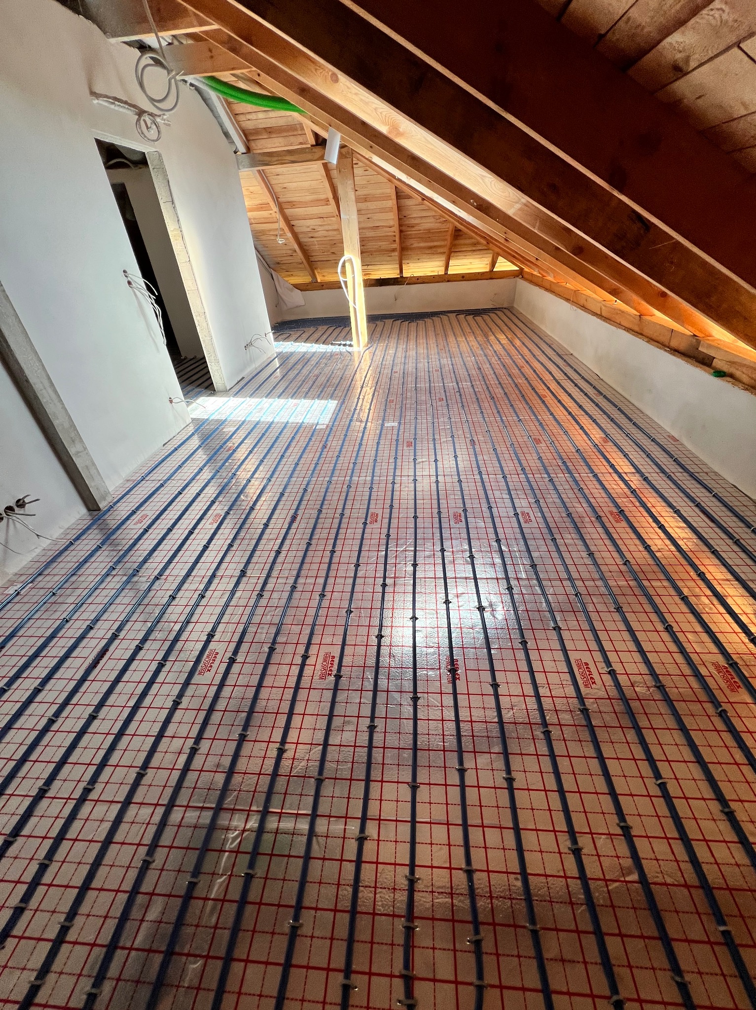 Instalacja ogrzewania podłogowego w poddaszu z widocznymi rurami grzewczymi ułożonymi na czerwonej siatce. Elementy instalacji przymocowane do podłoża. Widoczne drewniane elementy konstrukcji dachu.