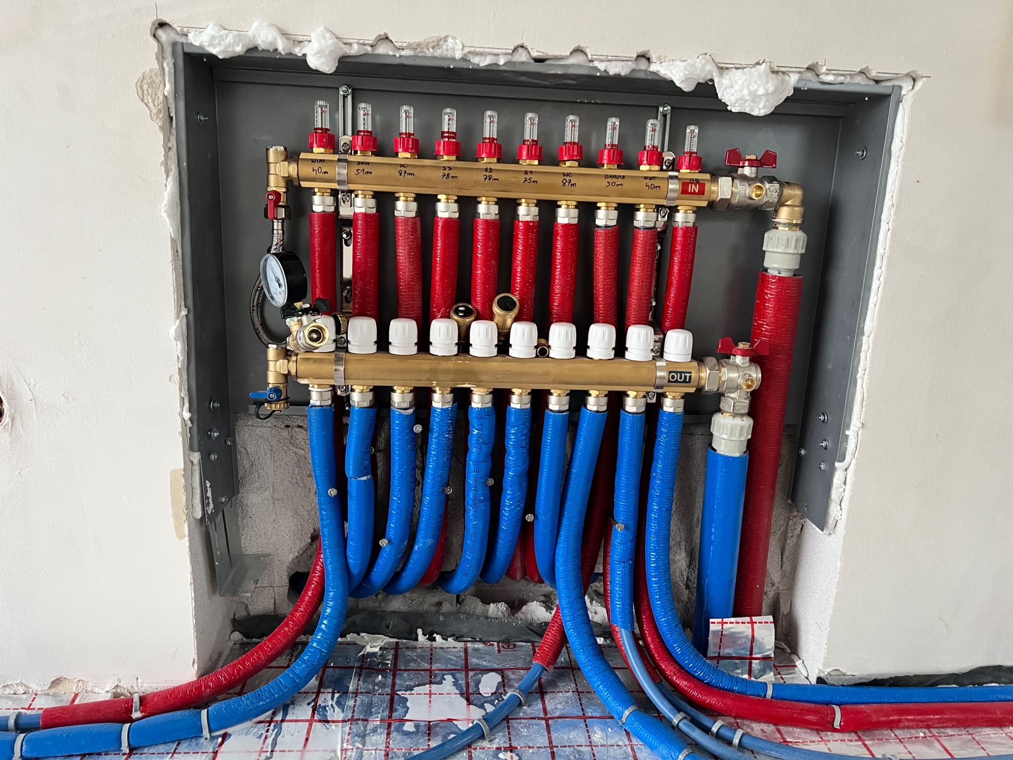 Rozdzielacz ogrzewania podłogowego z czerwono-niebieskimi rurami w szarej obudowie, widoczne oznaczenia długości pętli. Montaż w trakcie wykończenia.