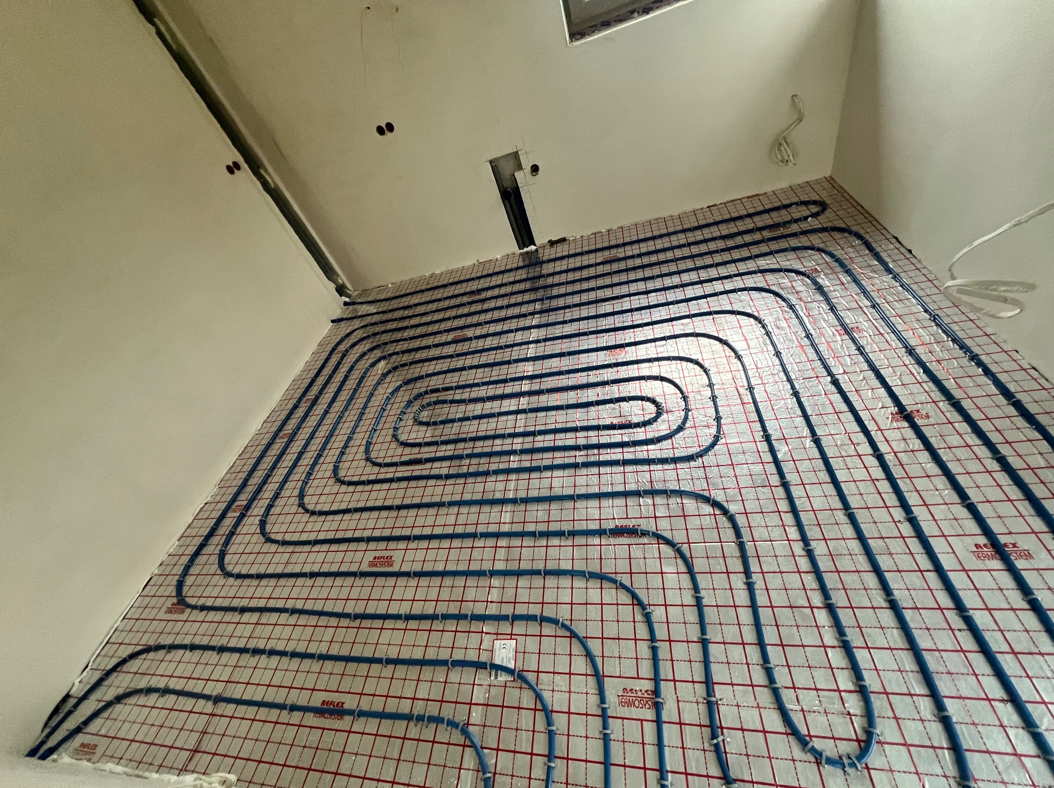 Błękitne rury ogrzewania podłogowego ułożone na izolacji z siatką w nowym budownictwie, gotowe do zalania wylewką. Widok z góry, surowy stan.